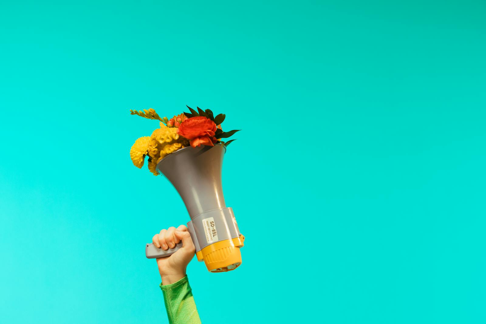 A creative composition featuring a hand holding a megaphone filled with vibrant flowers against a teal backdrop.